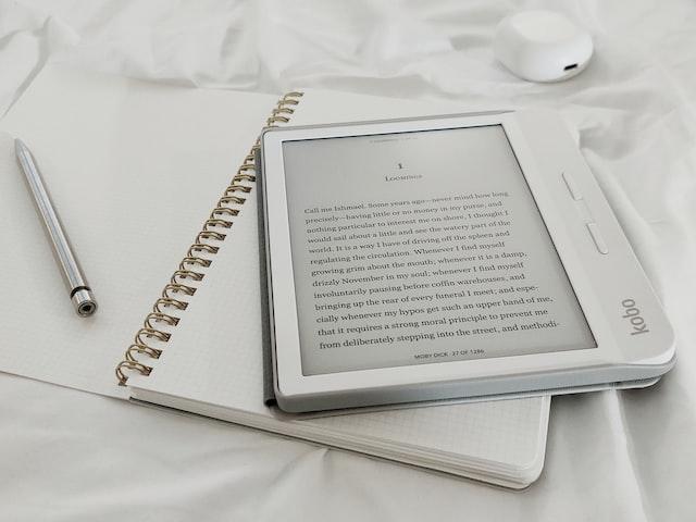 A white e-reader is laying on top of an open notebook with a pen to the left.