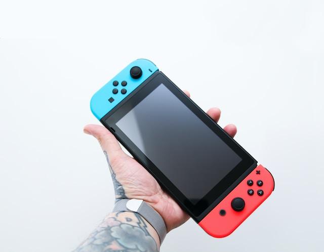 A light grey background with an extended tattoed arm and a hand holding a Nintendo Switch