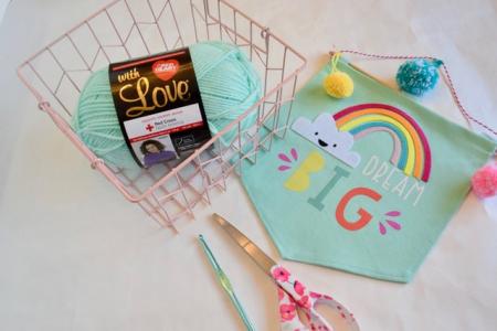 aqua yarn in a pink wire basket with scissors and a crochet needle to the side of the basket. There is a wall pennant with a rainbow with the words "Dream BIG" underneath.