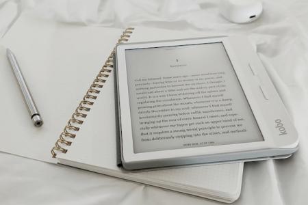A white e-reader is laying on top of an open notebook with a pen to the left. 