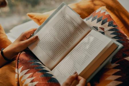 hands holding a book with a pillow and blanket on lap.